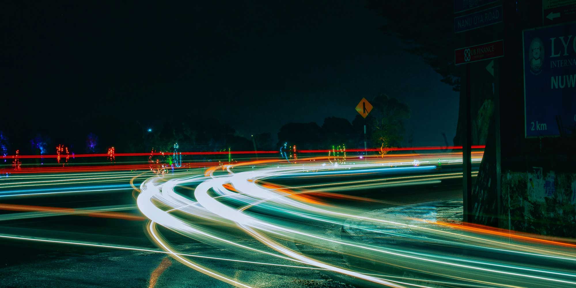 Nachtaufnahme einer Straßenkreuzung auf der nur die Lichter der vorbeifahrenden Autos als Lininen zu erkennen sind.